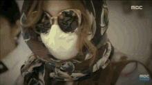 a woman wearing sunglasses and a scarf has a mbc logo on the bottom right