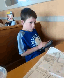 a young boy wearing a blue college shirt sits at a table using a tablet