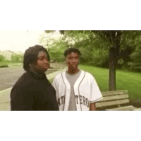 two men are standing next to each other in a park . one of the men is wearing a baseball uniform .
