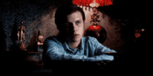 a man in a blue shirt is sitting at a table in a dark room with a lamp in the background .