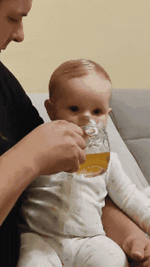 a baby is drinking from a glass while sitting on a man 's lap