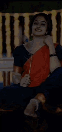 a woman in a red and blue saree is sitting on a couch holding a flower and smiling .