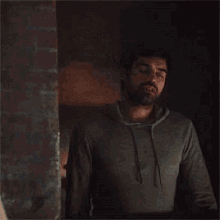 a man with a beard wearing a grey hoodie stands in front of a brick wall