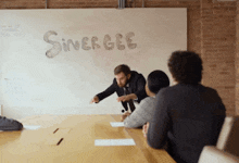 a group of people are sitting at a table in front of a white board that says synergee