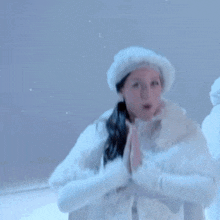 a woman in a white fur coat and hat smiles with her hands folded