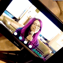 a woman with purple hair and glasses is holding a microphone in front of a blackmagic monitor