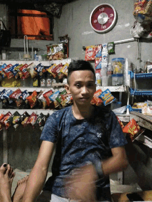 a man in a blue shirt is sitting in front of a shelf full of snacks and a clock that says 12 on it