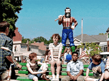 a group of children sit on a green bench in front of a statue of a man with a baseball bat