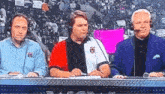 three men are sitting at a table with microphones in front of a crowd of people .