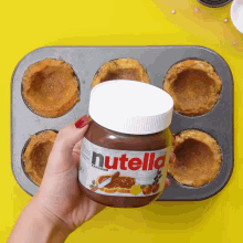 a person is holding a jar of nutella over a muffin tin