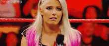 a woman with blonde hair and pink highlights is standing in a wrestling ring with a microphone in her hand .