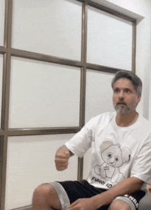 a man wearing a white t-shirt with a teddy bear on it is sitting in front of a window