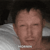 a man without a shirt is laying in bed with his eyes closed and the word mornin written on his face .