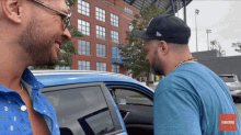 two men are talking in front of a blue car with a subscribe button on the bottom