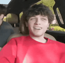a young man in a red sweater is sitting in the back seat of a car and smiling .
