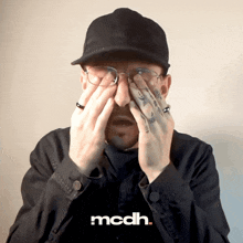 a man wearing glasses and a hat is covering his face with his hands and the word mcdh is on the bottom