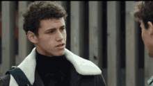 a man with curly hair wearing a black turtleneck and a white jacket