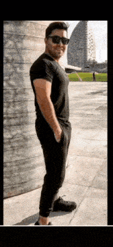 a man wearing sunglasses and a black shirt is standing on a sidewalk in front of a building .