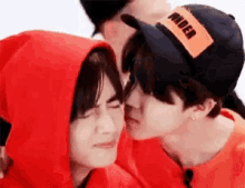 a man in a red hoodie is kissing another man in a hat .