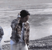a man in a camouflage jacket is standing on a rocky beach next to a body of water .