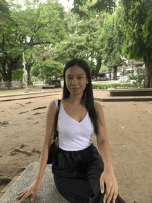a woman in a white tank top and black pants is sitting on a bench in a park