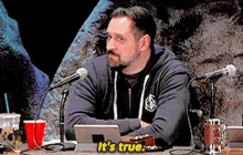 a man sitting at a table with two microphones and a sign that says " it 's true "