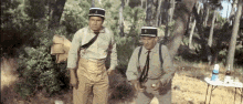 two men in military uniforms are standing in the woods .