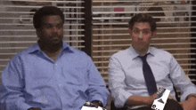 two men are sitting next to each other in front of a window in an office .