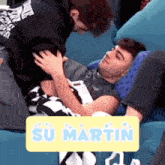 two men laying on a blue couch with the name su martin on the bottom right