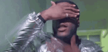 a man with a beard and a silver jacket is covering his eyes with his hand .