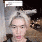 a young man with blonde hair is standing in front of a building and looking at the camera .