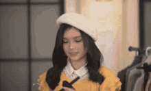 a woman wearing a white beret and a yellow shirt is looking at her phone .