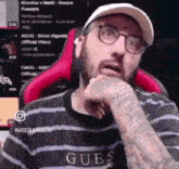 a man with a beard and glasses is wearing a hat and a guess shirt .