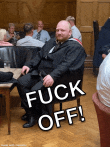 a man in a suit sits in a chair with the words " fuck off " written above him