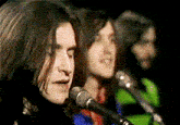a man with long hair singing into a microphone in front of two other men