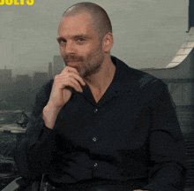 a man with a shaved head and a beard is sitting in front of a window with the word bolts on it