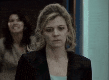a woman in a black jacket stands in a hallway with another woman behind her