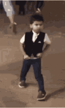 a young boy wearing a vest and a white shirt is standing on a sidewalk .