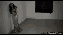 a woman in a long silver dress is standing in a dark room against a wall .