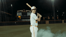 a baseball player is holding a bat in front of a scoreboard that says claude m smith field