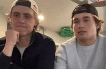 two young men are sitting next to each other in a room . one of the men is wearing a red bull hat .