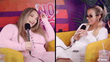 two women are sitting on a couch with microphones in front of them and one of them is wearing glasses .