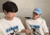 two young men are sitting next to each other on a couch . one of the men is wearing a supreme hat .