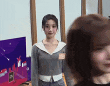 a woman in a gray sweater is standing next to another woman in front of a television .
