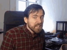 a man with a beard wearing a plaid shirt is making a funny face while sitting in a chair .