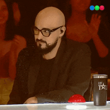 a man with glasses and a beard sits at a table with a cup that says got