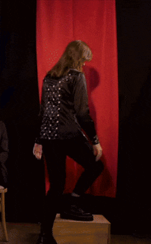 a woman in a black jacket and black pants is standing on a box in front of a red curtain