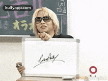 a man is holding a white board with a signature on it in front of a blackboard .
