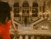 a woman in a red dress is standing on a balcony overlooking a staircase in a building .