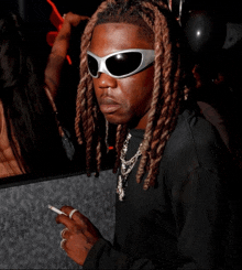 a man with dreadlocks wearing sunglasses holds a cigarette in his hand